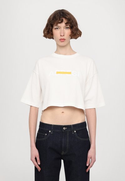 Fehér, cropped póló sárga és fehér grafikai szöveggel. Sötétkék farmernadrággal párosítva, kontrasztos varrással és fém kiegészítőkkel.