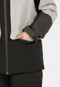 Veste imperméable avec un design bicolore en gris et noir, dotée d'une fermeture éclair à l'avant, de poignets réglables et de poches latérales.
