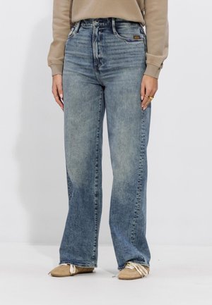 Mid-rise, lichtvervaagde blauwe wijde jeans gecombineerd met beige sneakers en een beige sweatshirt, gedragen door een staande persoon.