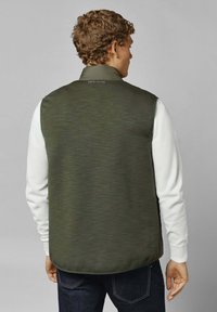 Donkergroen vest met een gestructureerd oppervlak, hoge kraag en logo-detail op de rug, gecombineerd met een witte langemouwen shirt en donkere jeans.