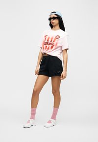 Ροζ γραφικό t-shirt με κείμενο «JUST DO IT», μαύρο σορτς, ροζ κάλτσες και λευκά αθλητικά παπούτσια με ροζ λεπτομέρειες. Ολοκληρώνεται με ανοιχτό μπλε καπέλο.