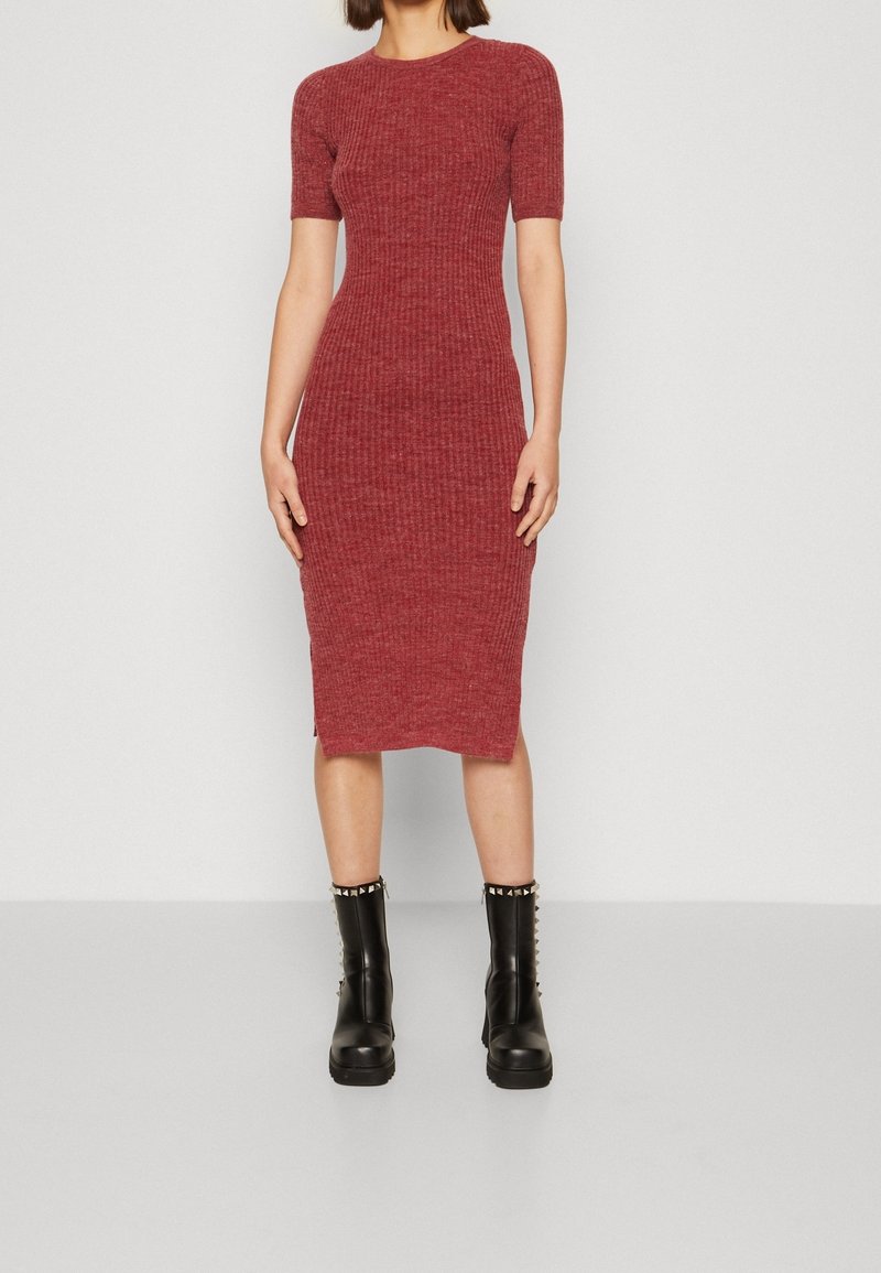 Femme portant une robe rouge ajustée, côtelée, longueur genou avec des manches courtes, associée à des bottines noires ornées de clous argentés.