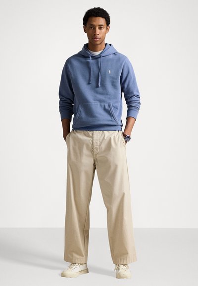Blauwe katoenen hoodie met een voorzak en een capuchon met koord, gecombineerd met beige baggy broeken en lichte sneakers, op een neutrale achtergrond.
