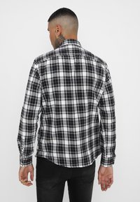 Chemise en flanelle à carreaux noir et blanc, avec un col rabattable et des manches longues. Le tissu a une texture douce. Vue de dos.