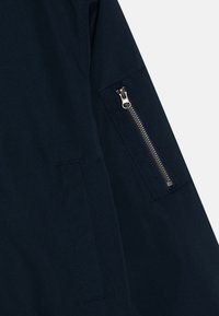 Manica di giacca blu navy con una tasca con zip visibile, dotata di hardware argento e texture liscia, insieme a un dettaglio di cucitura piatta.
