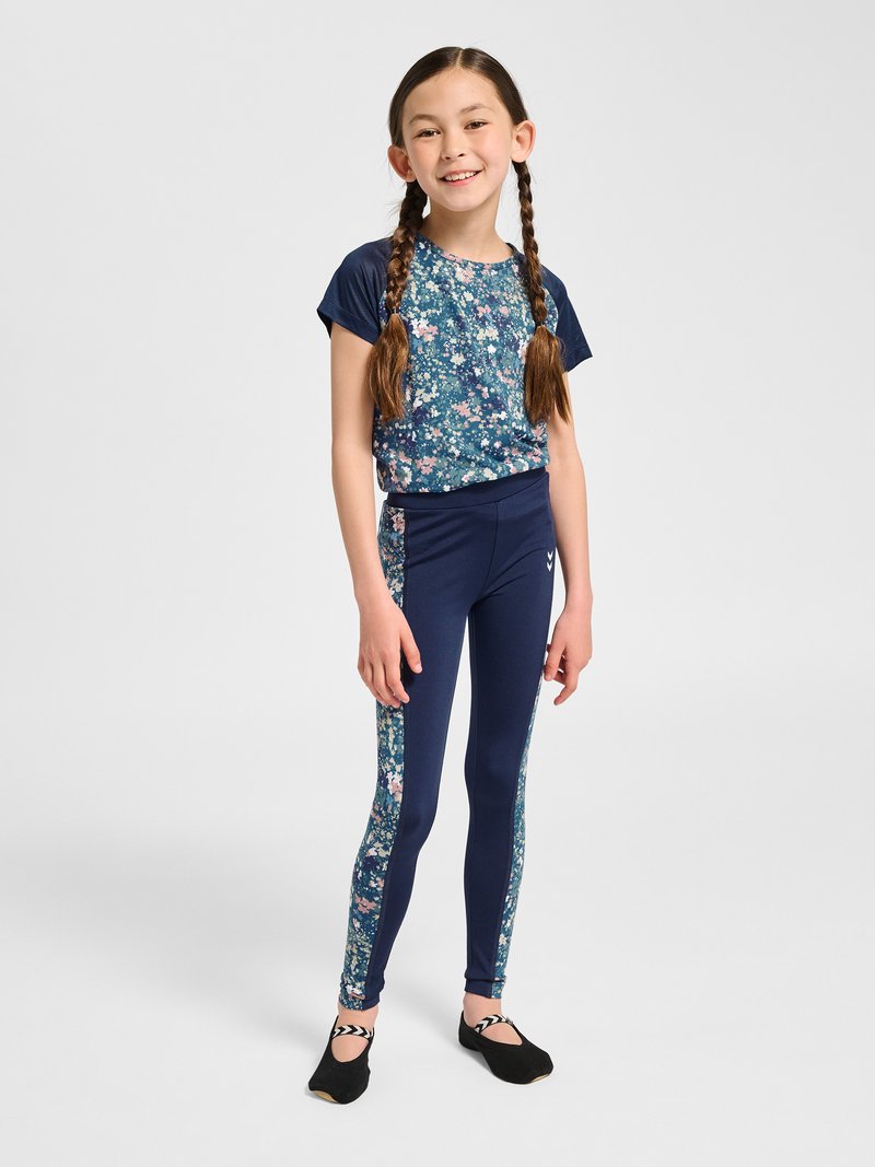 Jeune fille aux cheveux tressés portant une tenue de sport bleu marine à motifs floraux et des ballerines noires, debout et souriante sur fond uni.