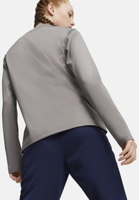 Giacca sportiva grigia con una superficie liscia e maniche lunghe, abbinata a pantaloni blu scuro. La giacca ha un taglio semplice e moderno.