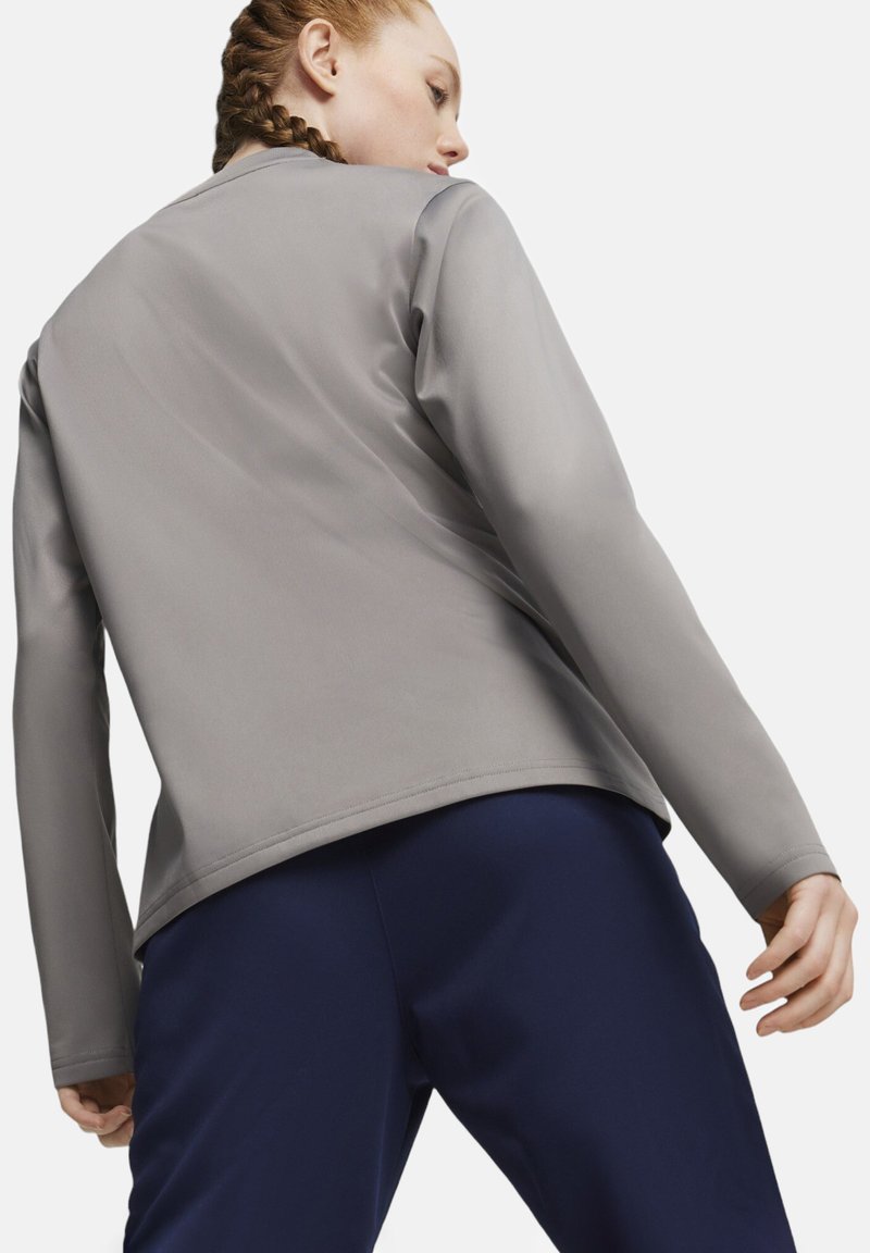Giacca sportiva grigia con una superficie liscia e maniche lunghe, abbinata a pantaloni blu scuro. La giacca ha un taglio semplice e moderno.