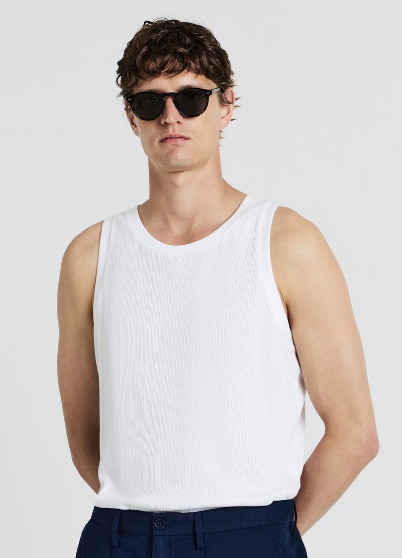 Débardeur sans manches blanc en tissu côtelé texturé. Modèle portant des lunettes de soleil rondes noires et un pantalon bleu marine. Design simple et ajusté.