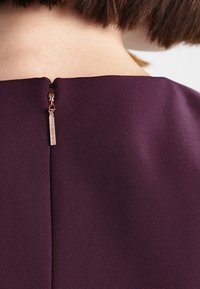 Detailní záběr na krk a horní část zad osoby, která má na sobě tmavě fialový oděv s růžově zlatým zipem na výstřihu.