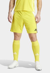 Geel sportieve shorts met zwarte accenten, gecombineerd met gele kniekousen en witte voetbalschoenen. Het materiaal lijkt lichtgewicht en glad.