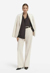 Weißer Blazer mit Nadelstreifen und passenden weiten Hosen über einer dunklen Bluse, akzentuiert mit einem schwarzen Gürtel. Schwarze spitz zulaufende High Heels vervollständigen den Look.