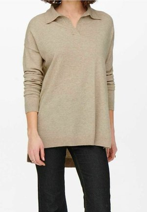 Pull-over en polo tricoté beige à manches longues, avec col et fentes latérales, associé à un jean foncé. Il présente une coupe décontractée et une texture douce.