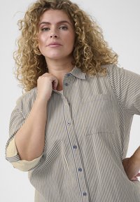 Chemise rayée en coton crème et marine, avec un devant boutonné, un col et une poche poitrine unique. Les manches retroussées mettent en valeur une coupe décontractée.