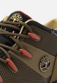 Ruskea kankainen kenkä, jossa on kuvioitu päällinen, punaiset nauhakujat ja kultainen Puu-logo sivussa. Varusteena pehmustettu kieli ja tummat yksityiskohdat.