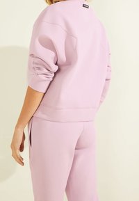 Pullover sweatshirt di un leggero lavanda con vestibilità rilassata, polsini a coste e un'etichetta sulla parte superiore della schiena, abbinato a pantaloni rilassati dello stesso colore.