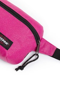 Sac à taille en tissu rose vif avec fermeture éclair noire, patch logo noir et sangle ajustable noire sur fond blanc.