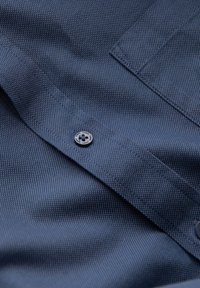 Camicetta blu navy con un tessuto testurizzato, caratterizzata da un piccolo bottone e un colletto piegato. Il tessuto presenta un sottile motivo a griglia.