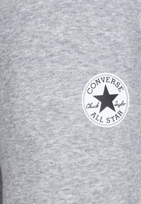 Grå hettegenser laget av mykt materiale med en sirkulær svart og hvit logo som har teksten "Converse All Star" og et stjernedesign.