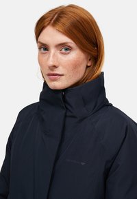 Roodharige vrouw met sproeten, gekleed in een donkerblauwe jas met hoge kraag, kijkt iets naar links tegen een effen lichte achtergrond.
