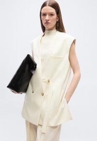 Γυναίκα με ίσια καστανά μαλλιά, φοράει αμάνικο crème σακάκι και παντελόνι σετ, κρατώντας μεγάλη μαύρη τσάντα χειρός, με το ένα χέρι στην τσέπη.