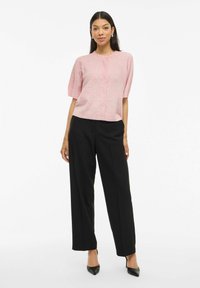 Cardigan rosa chiaro a maniche corte con scollatura rotonda e chiusura a bottoni, abbinato a pantaloni neri a gamba larga e tacchi neri.