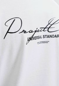 Valkoinen puuvillapaita, jossa on mustalla painettuna tekstiä, mukaan lukien "Project" ja "Universal Standard", sileä rakenne ja rento istuvuus.