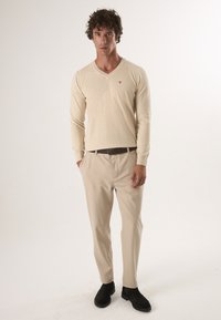 Beżowy sweter w serek z miękkiej przędzy, zestawiony z jasnobeżowymi spodniami. Czarne buty dopełniają outfit. Minimalne detale na swetrze.