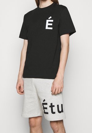 Personne portant un T-shirt noir avec un "É" blanc sur la poitrine et un short gris clair avec un grand texte noir "Étu" sur la jambe gauche.