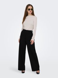 Top de manga larga beige, pantalones de pierna ancha de talle alto en negro y gafas de sol con tintado verde. Zapatos de tacón alto negros completan el conjunto.