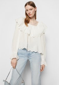 Blusa branca com camadas de babados, padrão de ilhós e mangas à boca de sino. Combinada com jeans azul claro e uma pequena mala cinza.