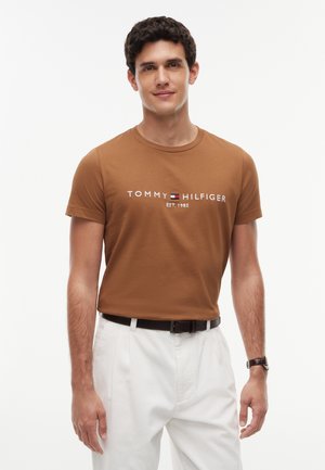Jeune homme portant un t-shirt marron Tommy Hilfiger et un pantalon blanc, debout devant un fond blanc uni.