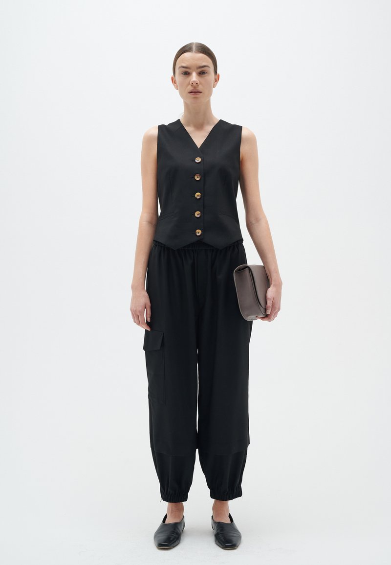 Schwarzes ärmelloses Oberteil mit fünf Holzknöpfen, kombiniert mit lockeren schwarzen Hosen mit elastischen Bündchen und Taschen. Hält eine braune Clutch.