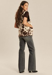 Strickweste mit einem beige-braunen Kuhmuster, relaxter Passform, mit Seitentaschen, getragen über einem kurzärmeligen braunen Oberteil und grauen Jeans.
