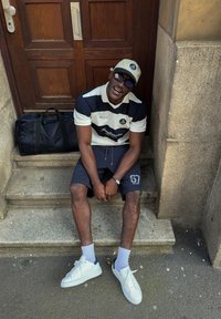 Homme portant des lunettes de soleil, une casquette, un polo rayé et un short, assis sur des marches en pierre près d'une porte en bois avec un sac de sport noir à côté de lui.