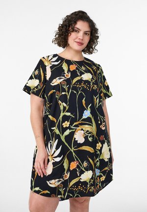 Žena s kučeravými vlasmi, ktorá nosí čierne šaty s krátkym rukávom, zdobené veľkými farebnými kvetmi a zelenými stonkami, stojí na bielom pozadí.