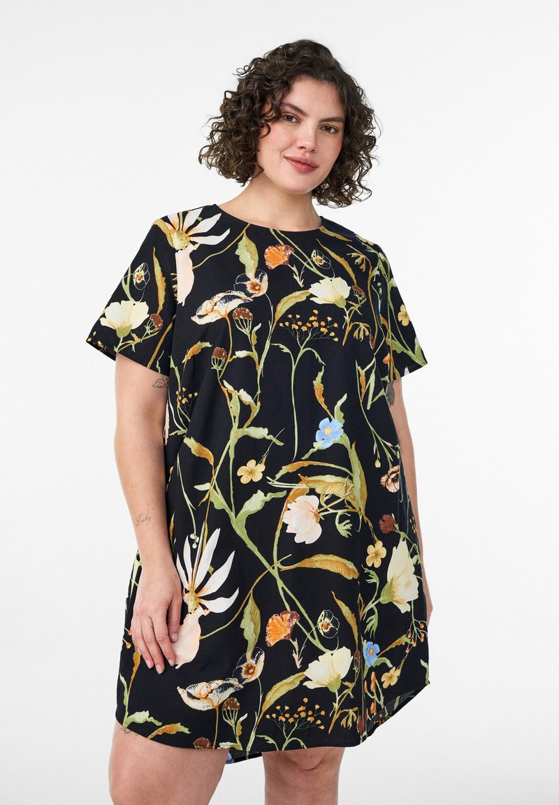 Žena s kučeravými vlasmi, ktorá nosí čierne šaty s krátkym rukávom, zdobené veľkými farebnými kvetmi a zelenými stonkami, stojí na bielom pozadí.