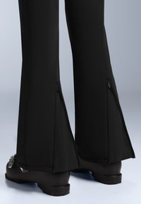 Fekete, harangszárú nadrág bokazippzárral, sima anyaggal és szoros szabással, sötét, lapos sarkú csizmával párosítva.
