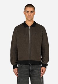 Rjava rebrasta jakna z zipom in črnim ovratnikom ter manšetami, ki se ponaša z drzno vertikalno črtasto zasnovo in zapiranjem na zadrgo. Teksture tkanine in minimalno blagovno znamko.