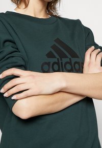 Sudadera verde de algodón con cuello redondo y mangas largas. Presenta un gran logo negro de Adidas en la parte frontal.