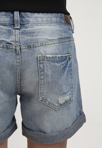 Pantalones cortos de mezclilla en azul claro con un bolsillo trasero desgastado, dobladillos doblados y una etiqueta de cuero en la cintura. Diseño clásico de cinco bolsillos.
