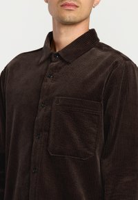 Brun fløjlsskjorte med krave, lodret ribbet tekstur og en enkelt brystlomme. Har sorte knapper og en afslappet pasform.