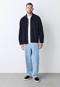 Schwarze Cordjacke mit Fronttasche, darunter ein weißes T-Shirt, hellblaue weit geschnittene Jeans und schwarze Sneaker. Einfaches und lässiges Outfit.