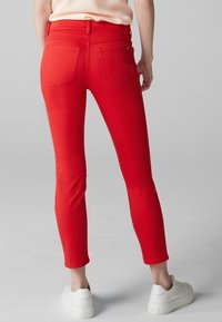 Jean stretch rouge à coupe slim, dotés de poches arrière et d'une taille mi-haute, associés à des baskets blanches sur un fond gris.