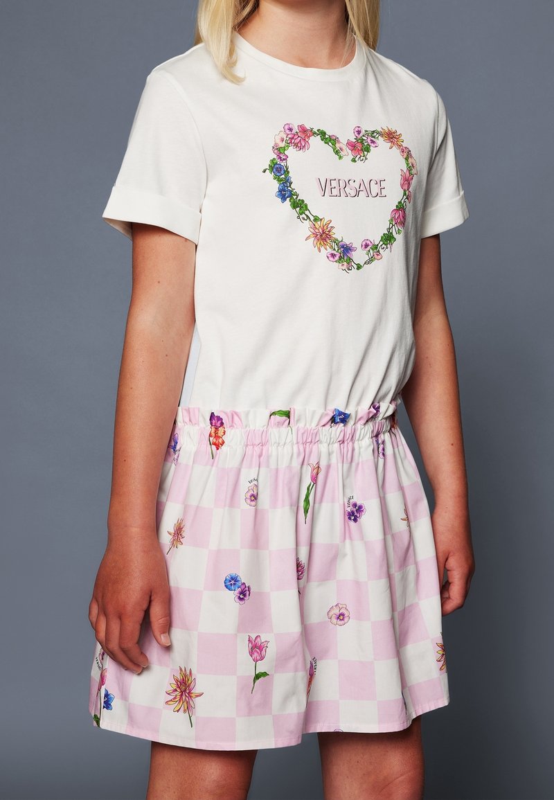 T-shirt blanc avec un motif en forme de cœur floral et le texte "VERSACE", associé à une jupe rose à carreaux avec des imprimés floraux multicolores et une taille élastique.