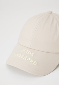 Beige cap made of textured fabric featuring embroidered "MADS NØRGAARD" in light green. Curved brim and stitched seams are visible.