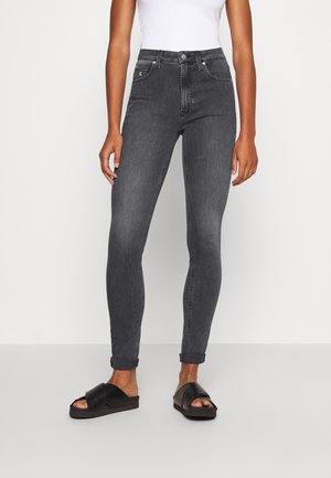 Femme portant un jean skinny gris foncé taille haute retroussé aux chevilles, un haut blanc sans manches et des sandales noires à enfiler.