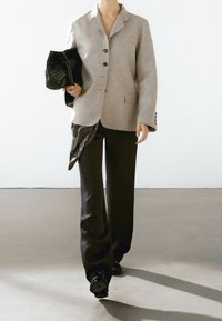 Blazer gris clair avec col cranté et trois boutons, associé à un pantalon large sombre. Sac à main noir tressé et détail d'écharpe à motifs.