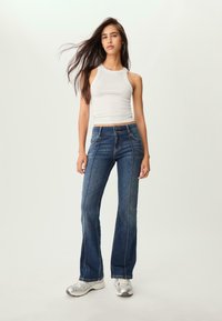 Jeansy dżinsowe w kolorze niebieskim z rozszerzającymi się nogawkami i wysokim stanem, z przednimi kieszeniami i kontrastowymi przeszyciami. W komplecie z białym topem bez rękawów i srebrnymi sneakersami.