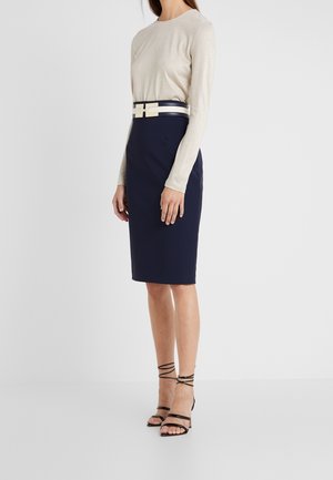 Femme portant un haut beige à manches longues, une jupe crayon bleu marine avec une large ceinture noire et blanche, et des sandales noires à talons hauts avec des lanières.